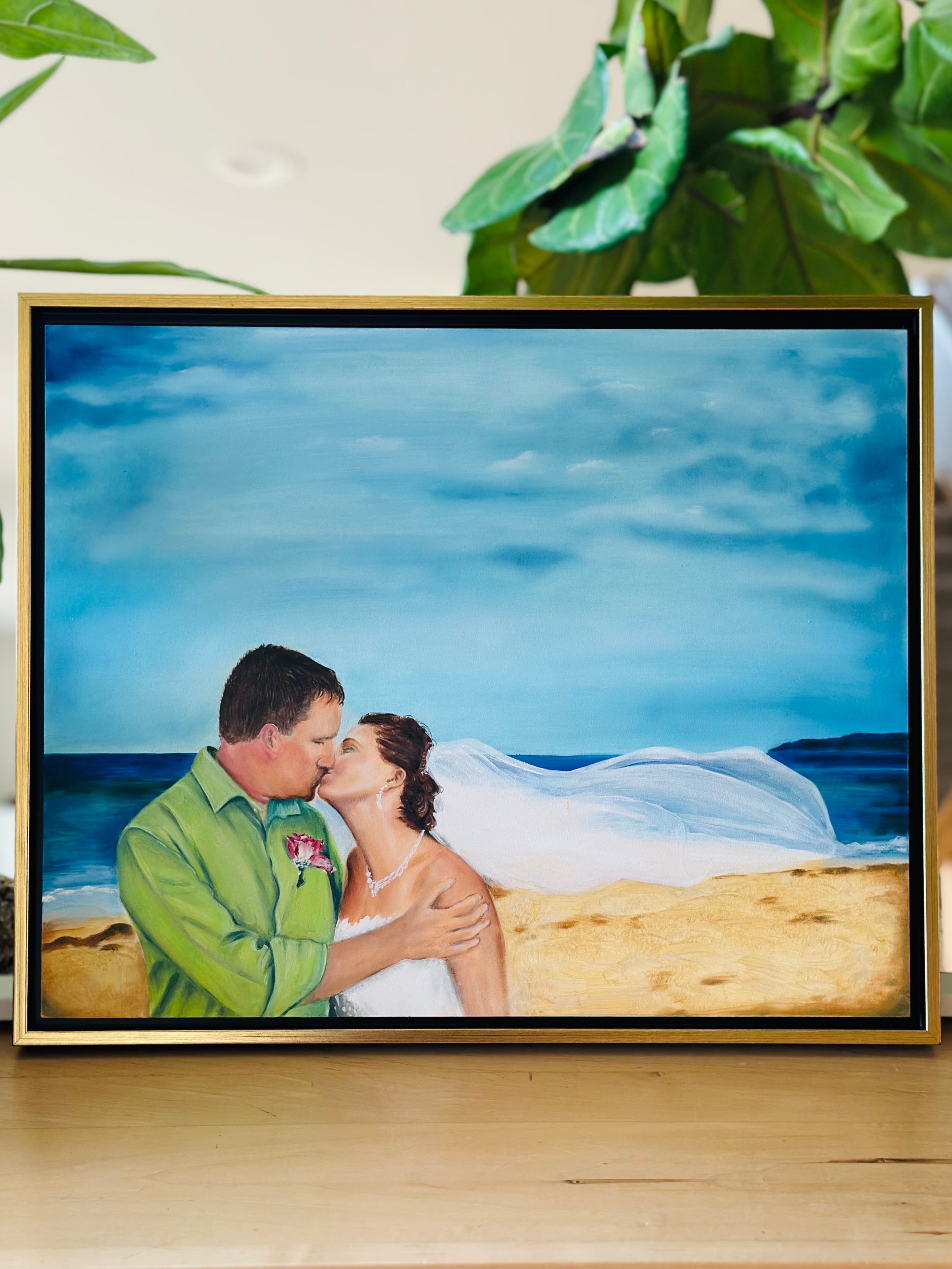 Seashore wedding painting — the couple kissing on the beach beneath a wide blue sky, in a gold frame