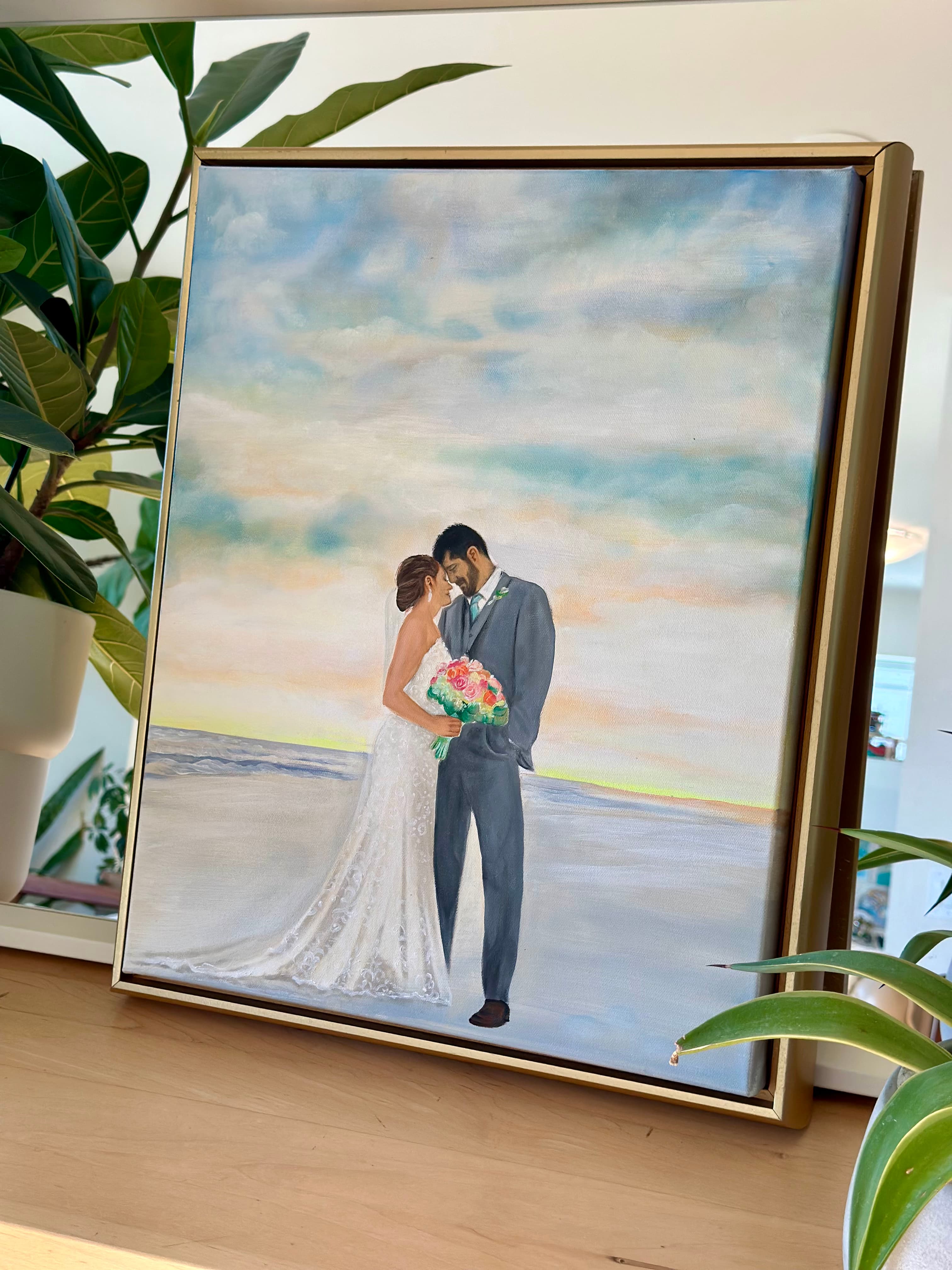 The beach sunset wedding painting displayed in its gold frame beside the studio plants
